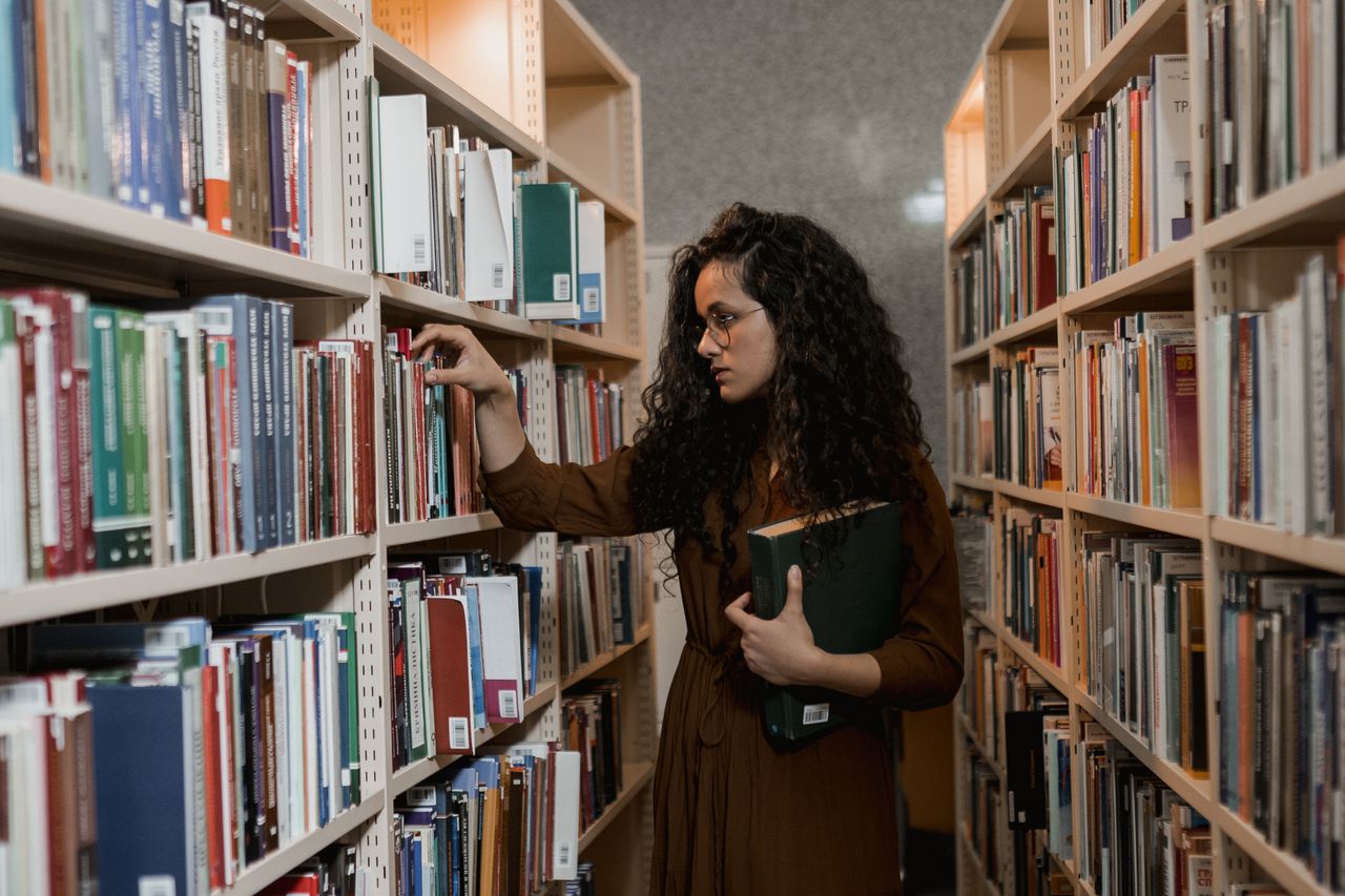 A woman with curly hair and eyeglasses choosing books in a library aisle filled with bookshelves.